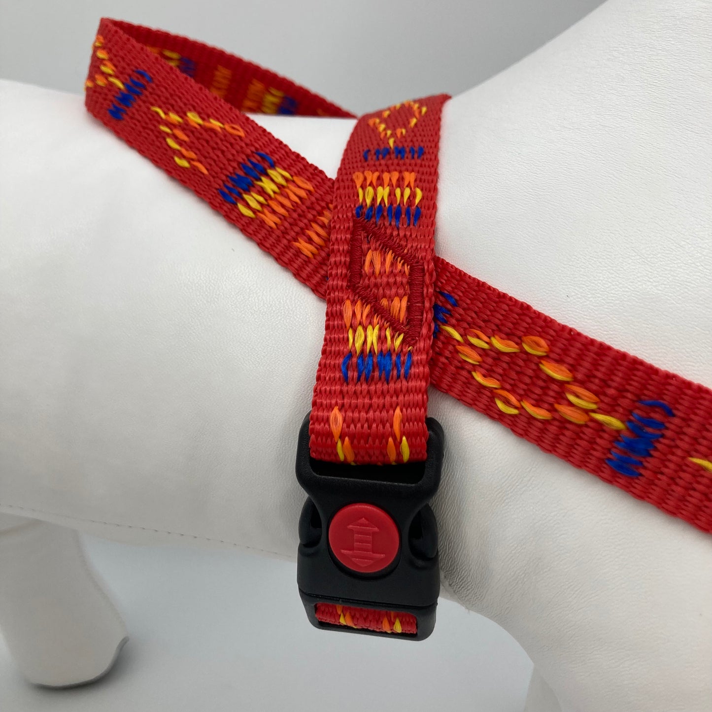 red Stitch (red fabric with print in 3 colors, yellow, orange and blue) harness displayed on a stuffed toy (dog), around the torso, showing the black lock and the loop on the back